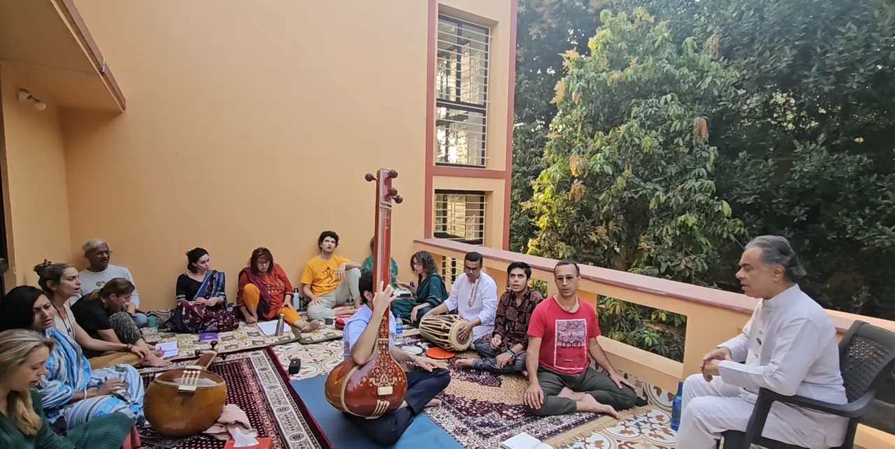 Participants seated on rugs on a terrace around a tanpura and a drum, listening to a man in white seated in a chair, with leafy trees beyond