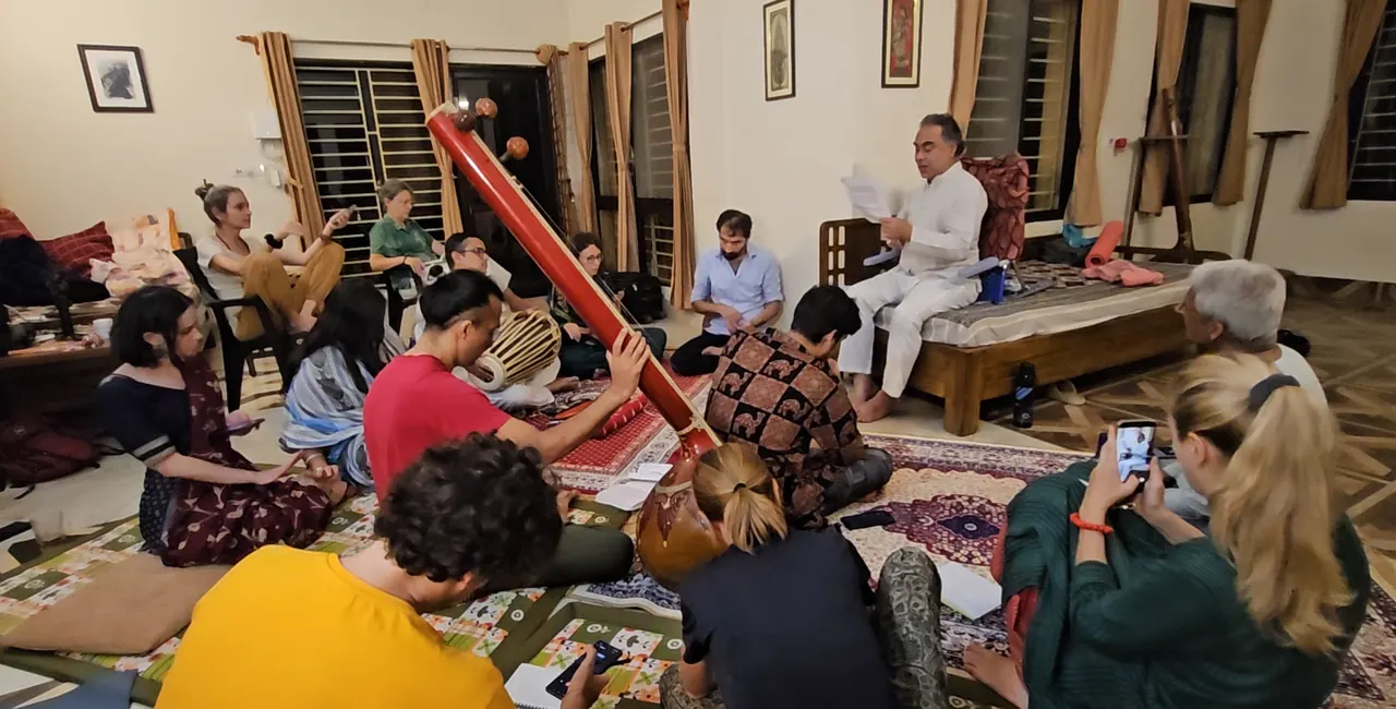 Participants seated on the floor of a room around a large tanpura, listening to a man in white who sits on a bed holding papers