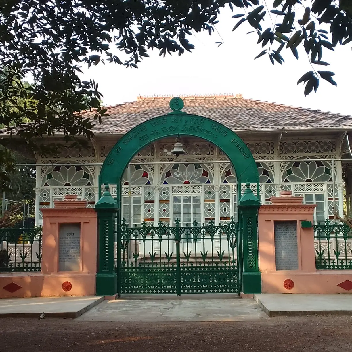 Ornate green iron arch and gate in front of a glass-windowed building with a tiled roof, framed by tree branches
