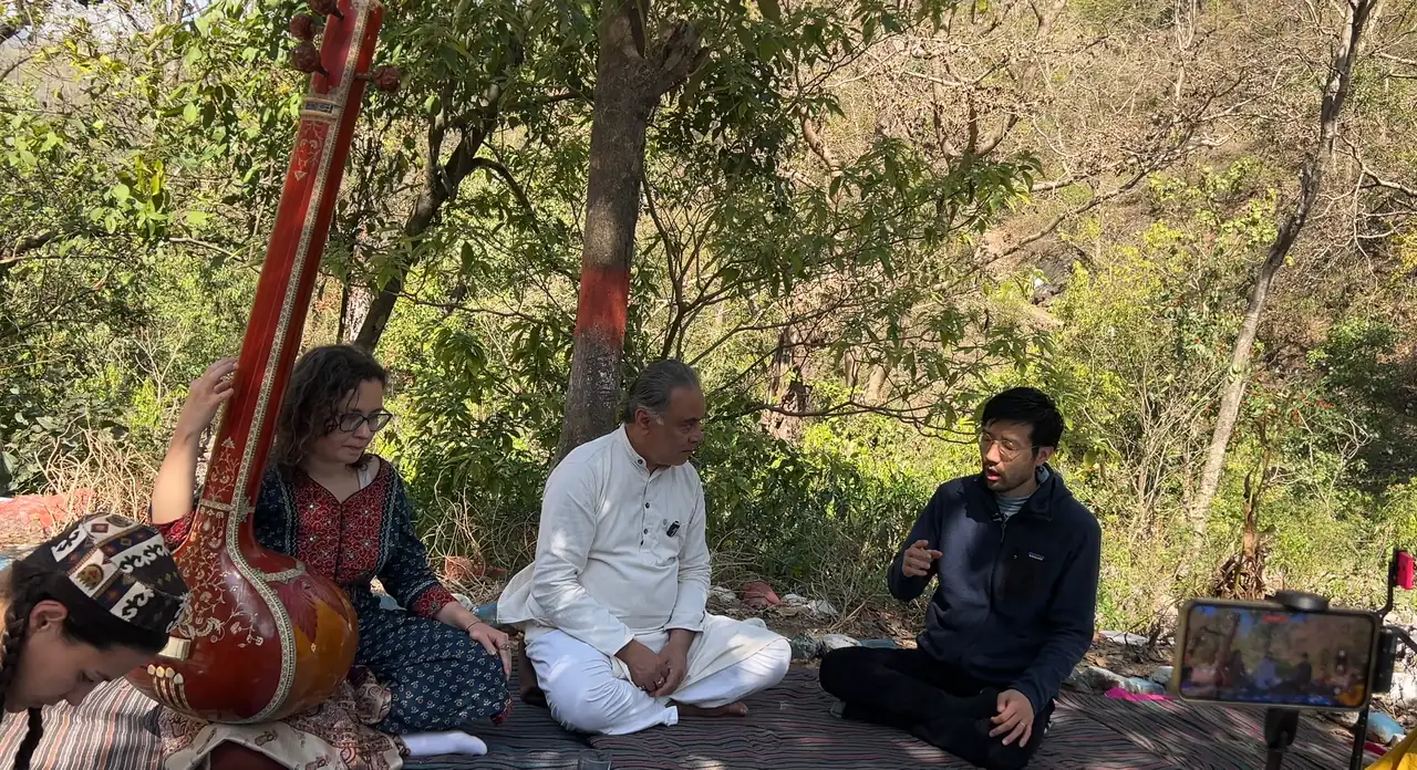 A man in white sits cross-legged on a mat under trees talking with a man in a dark jacket, while a woman holds a tanpura and a phone on a stand records