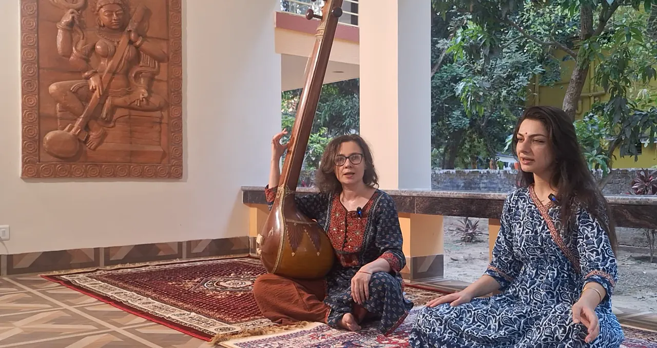 Two women sit cross-legged on rugs on a veranda, one holding a tanpura, in front of a carved wooden wall relief of a seated figure with a stringed instrument